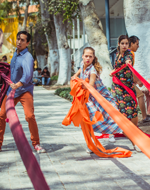 Image: Three dancers pull on brightly-colored fabric strips on the sidewalk in an urban setting.