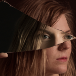 Image Detail: Close-up of a woman's face, which is turned towards the right and obscured by a shard of glass she is holding. The glass reflects a pair of eyes back, presumably the same woman's. She is dramatically lit against a black background.