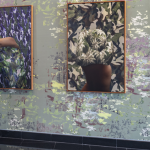 Four large collages hang in a gallery on top of a wallpaper. A person observes the installation as they walk by.