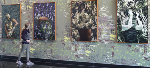 Four large collages hang in a gallery on top of a wallpaper. A person observes the installation as they walk by.