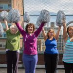 Six older women cheerleaders dressed colorfully with their silver pom-poms raised above their heads as they cheer from the rooftop of the 14th Street Y on a sunny day.